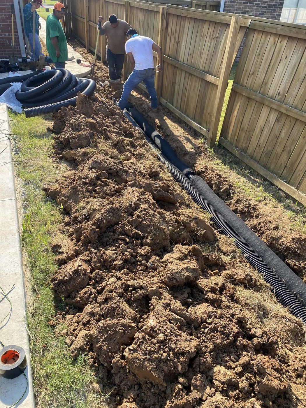 Crew laying corrugated drainage pipe in an open trench along a wood privacy fence